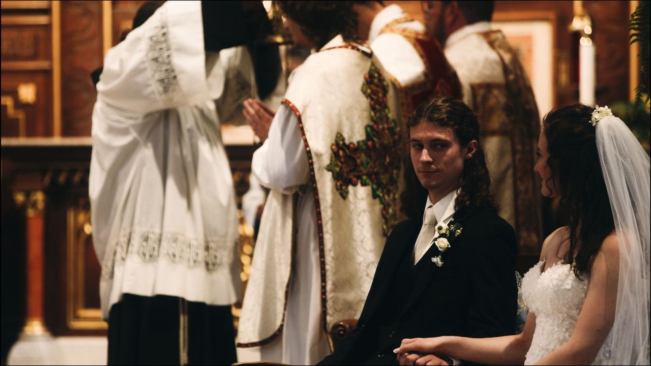 Bride and groom sitting in sanctuary while clergy perform mass in background.