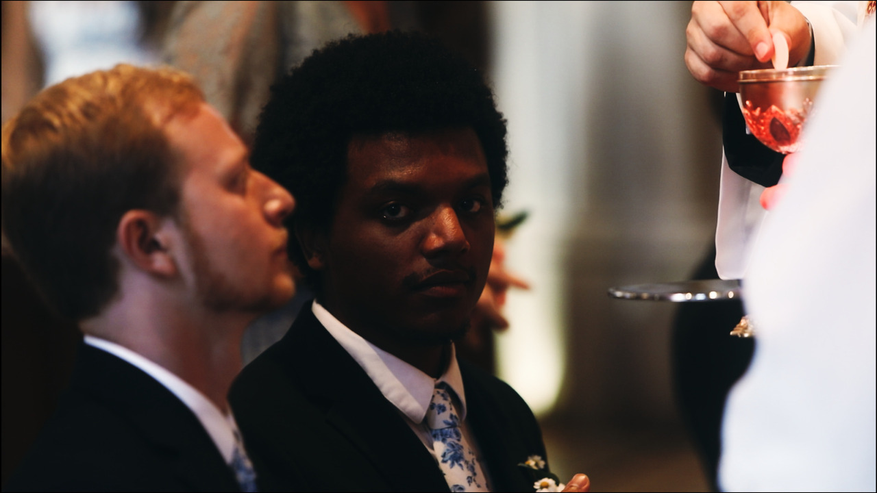 Priest handing sacred host to groomsman while second groomsman looks at camera.
