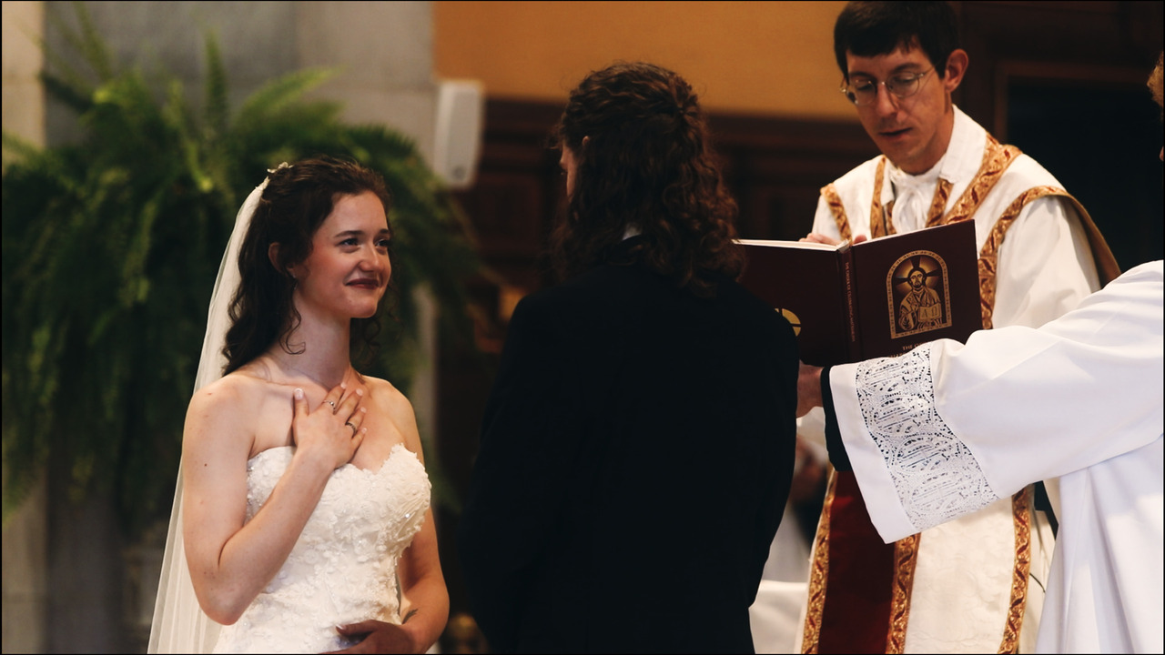 Bride smiles at groom while priest prays over them.
