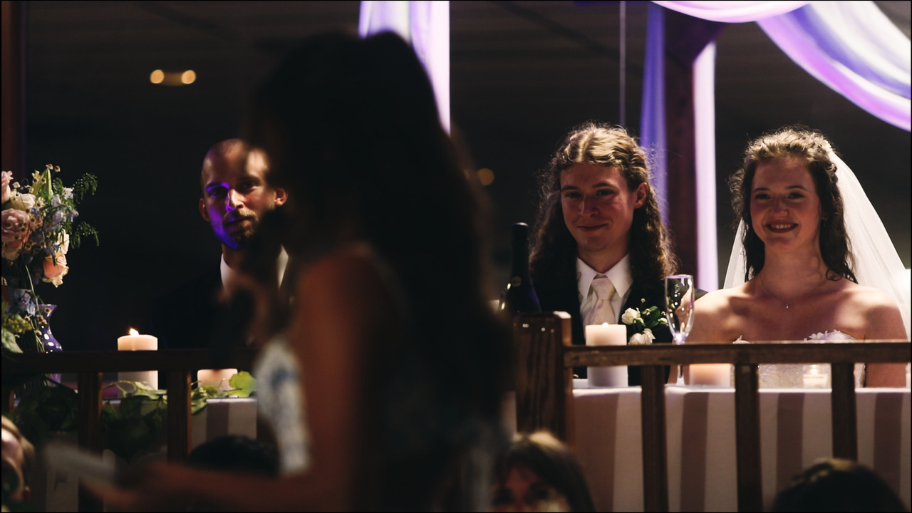 Bride and groom sitting, watching bridesmaid toast in foreground.