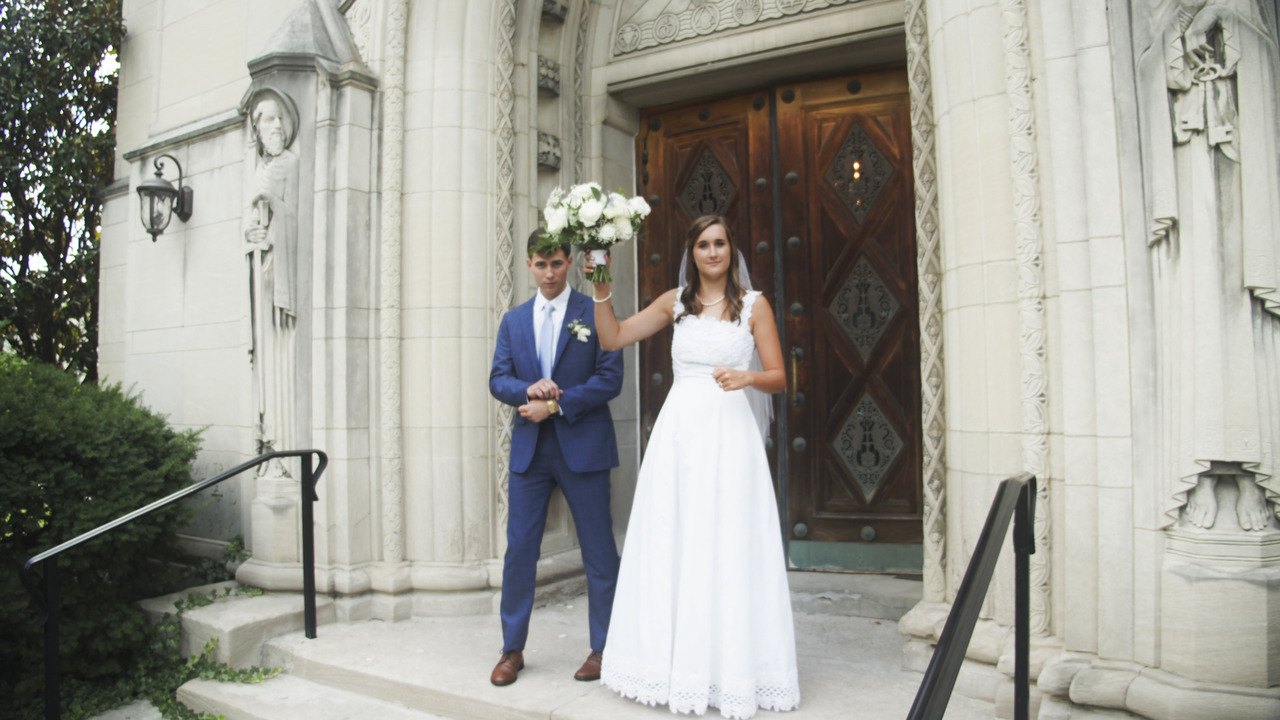 Bride and groom stand on church steps, bride waves bouquet at camera.