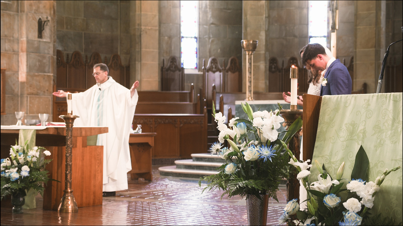 Bride and goom stand and pray to side of alter while priest prays behing it.