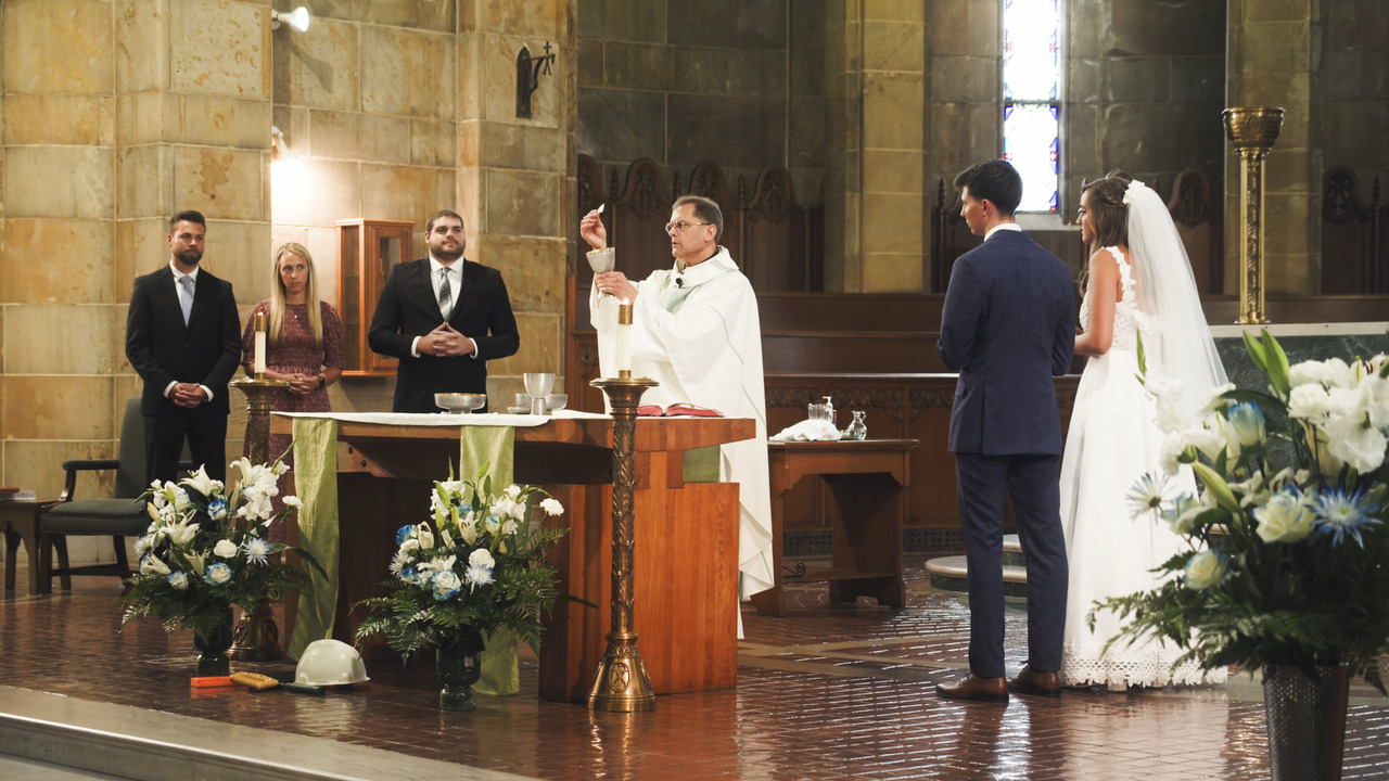 Priest holds sacred host aloft while Bride and Groom stand to side of altar and watch.