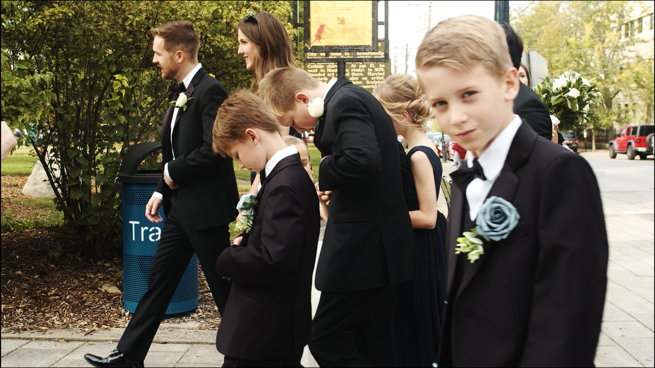 Boy looking at camera while he and rest of groomsmen walk down sidewalk.
