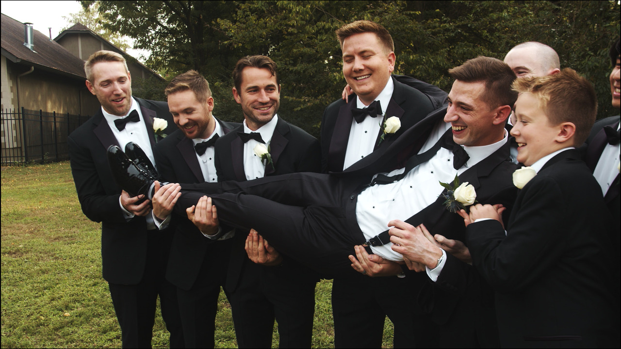 Groomsmen hold groom in their arms and laugh for group photo.