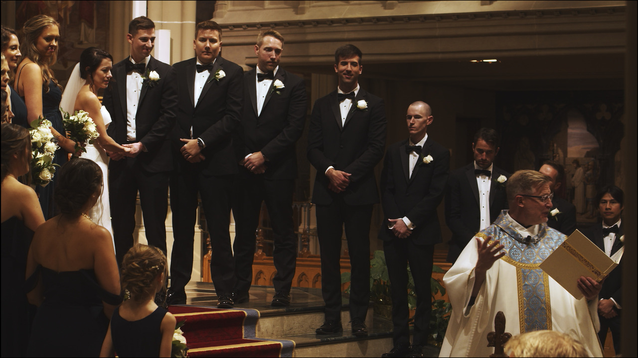 Bride and groom holding hands in front of altar, flanked on both sides by bridal party, watching priest give homily at bottom of steps.