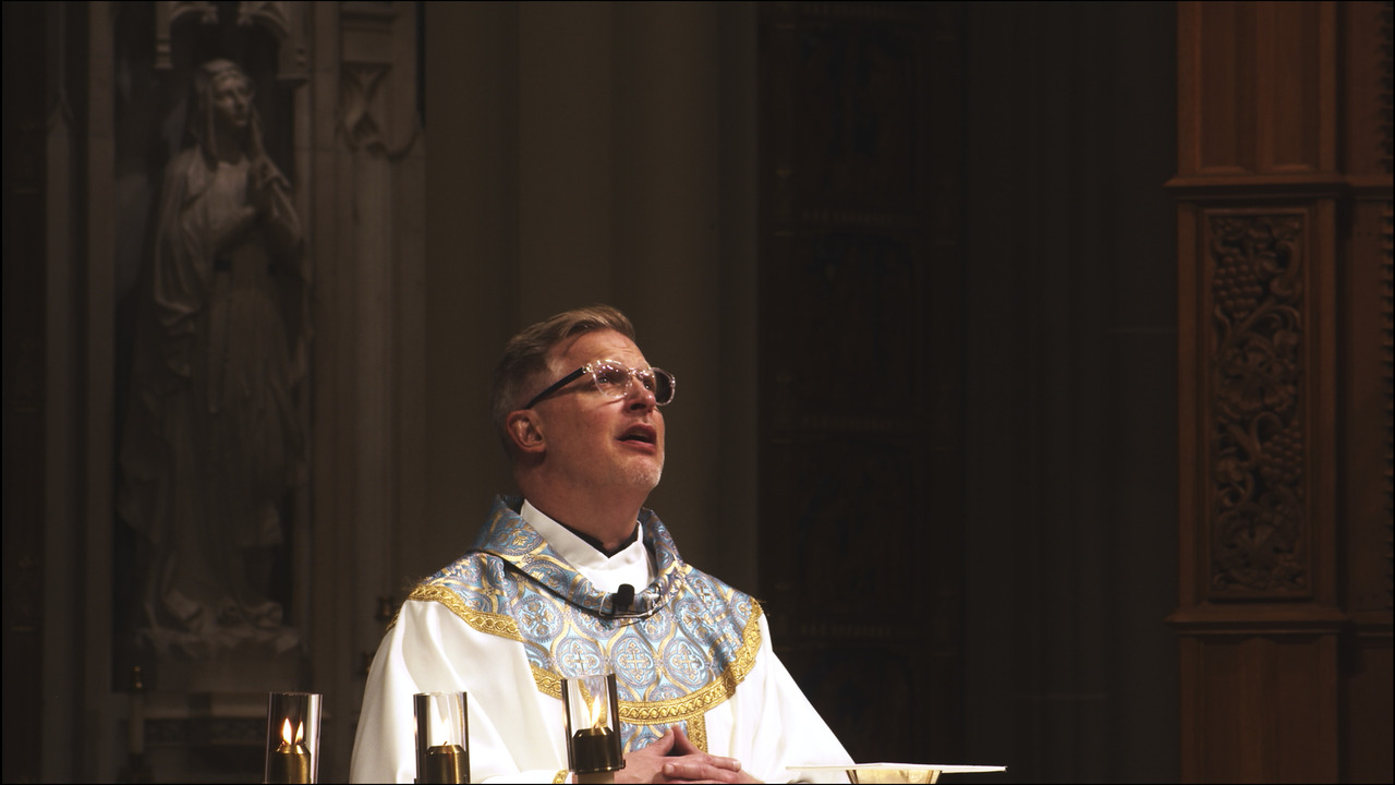 Priest looking up and praying, with statue of Virgin Mary behind him.