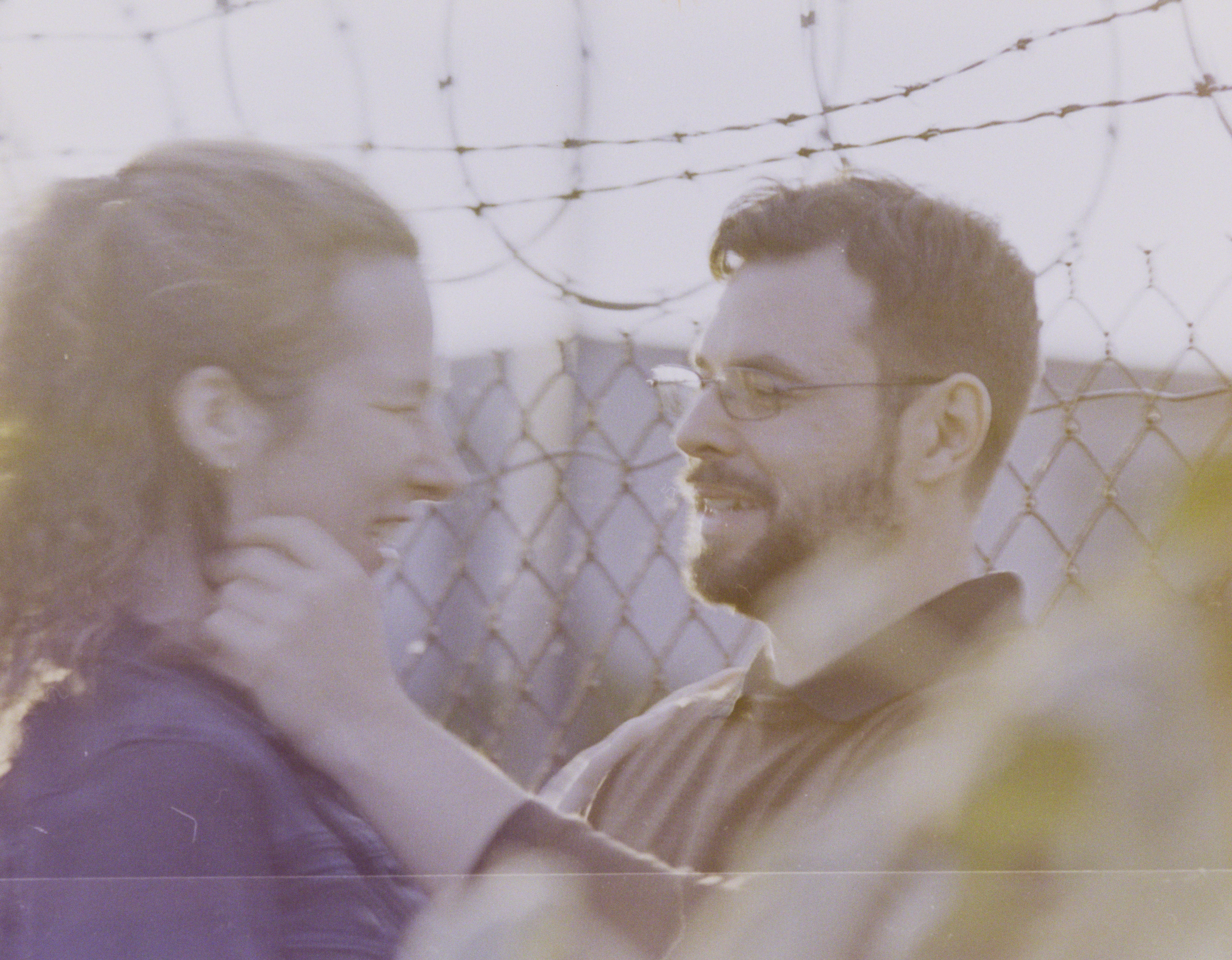 Couple smiling and staring into each other's eyes in front of razorwire fence, color.